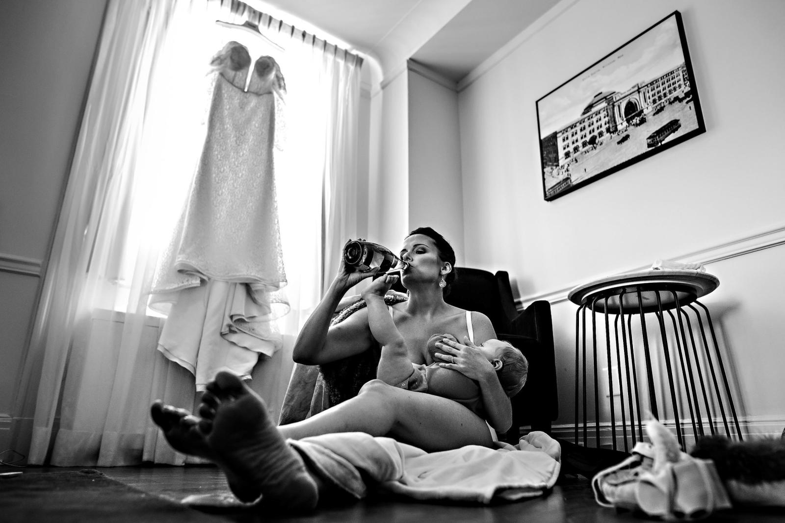 Bride breast-feeding and drinking from a wine bottle on the floor with wedding dress hung in the window