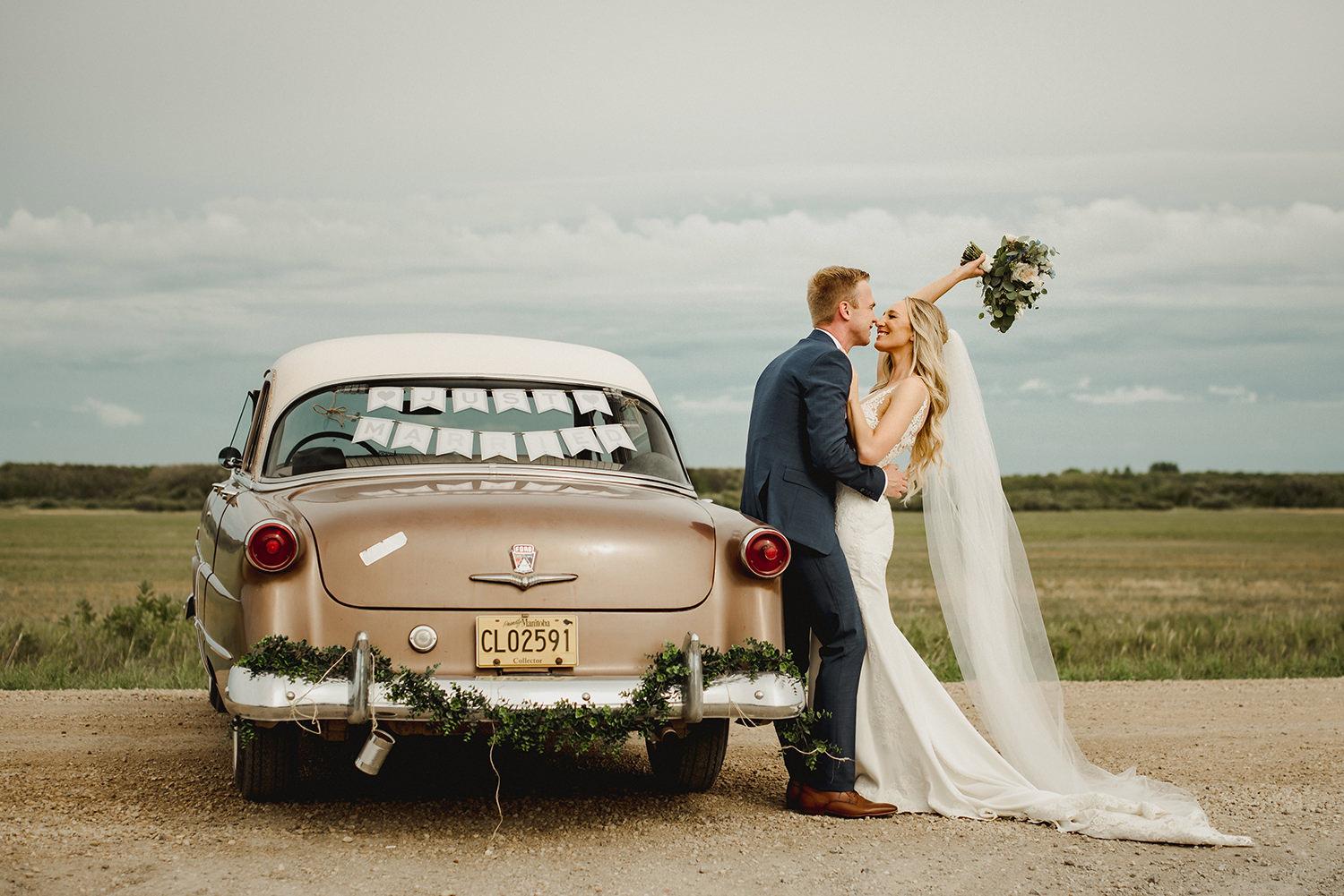 Bride and groom kissing in front of classic car with 'Just Married' sign at Hawthorn Estates in Manitoba